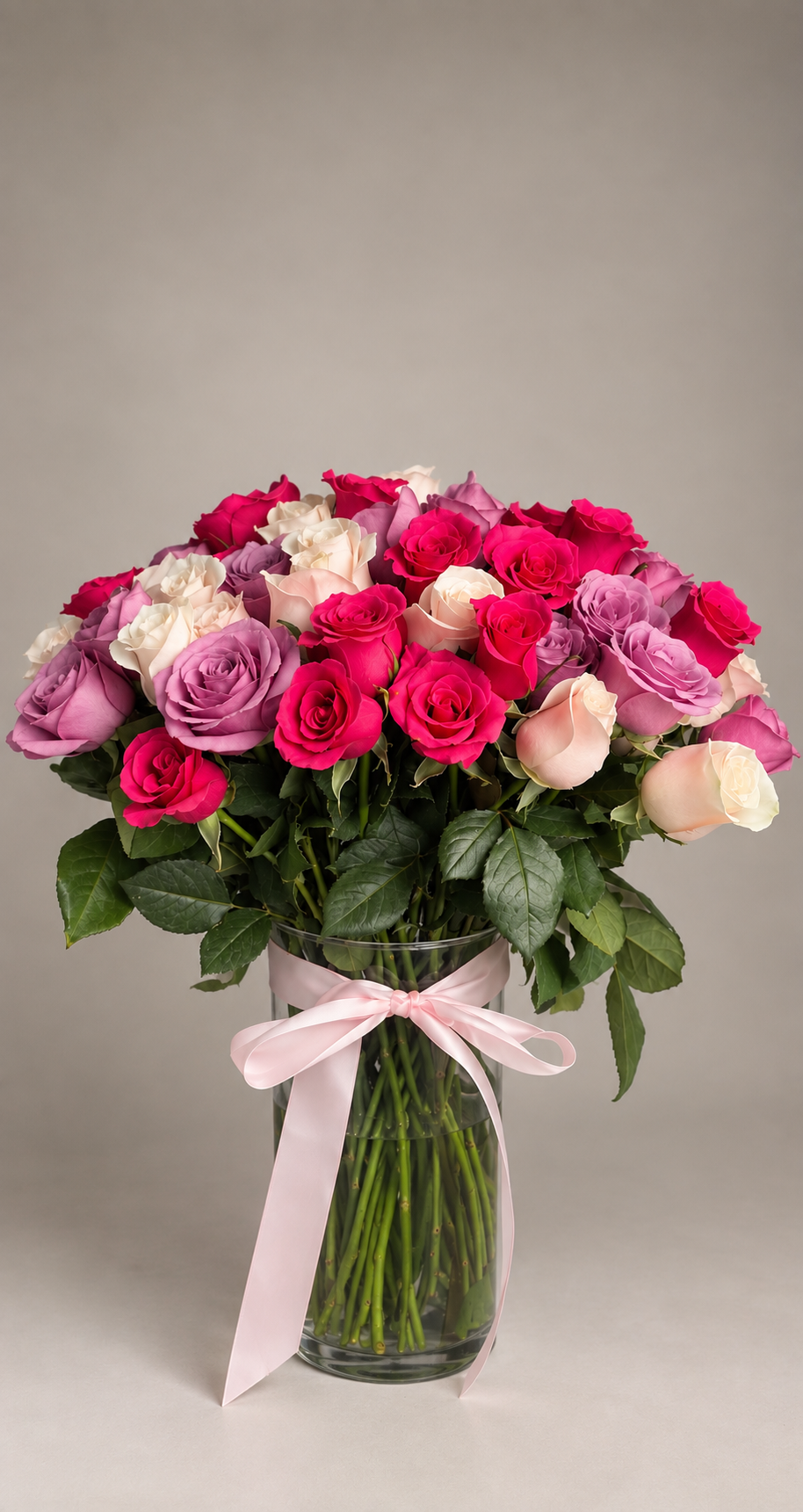 Florero de cristal con rosas en tonos fucsia, lila y marfil, rematado con un lazo rosa.