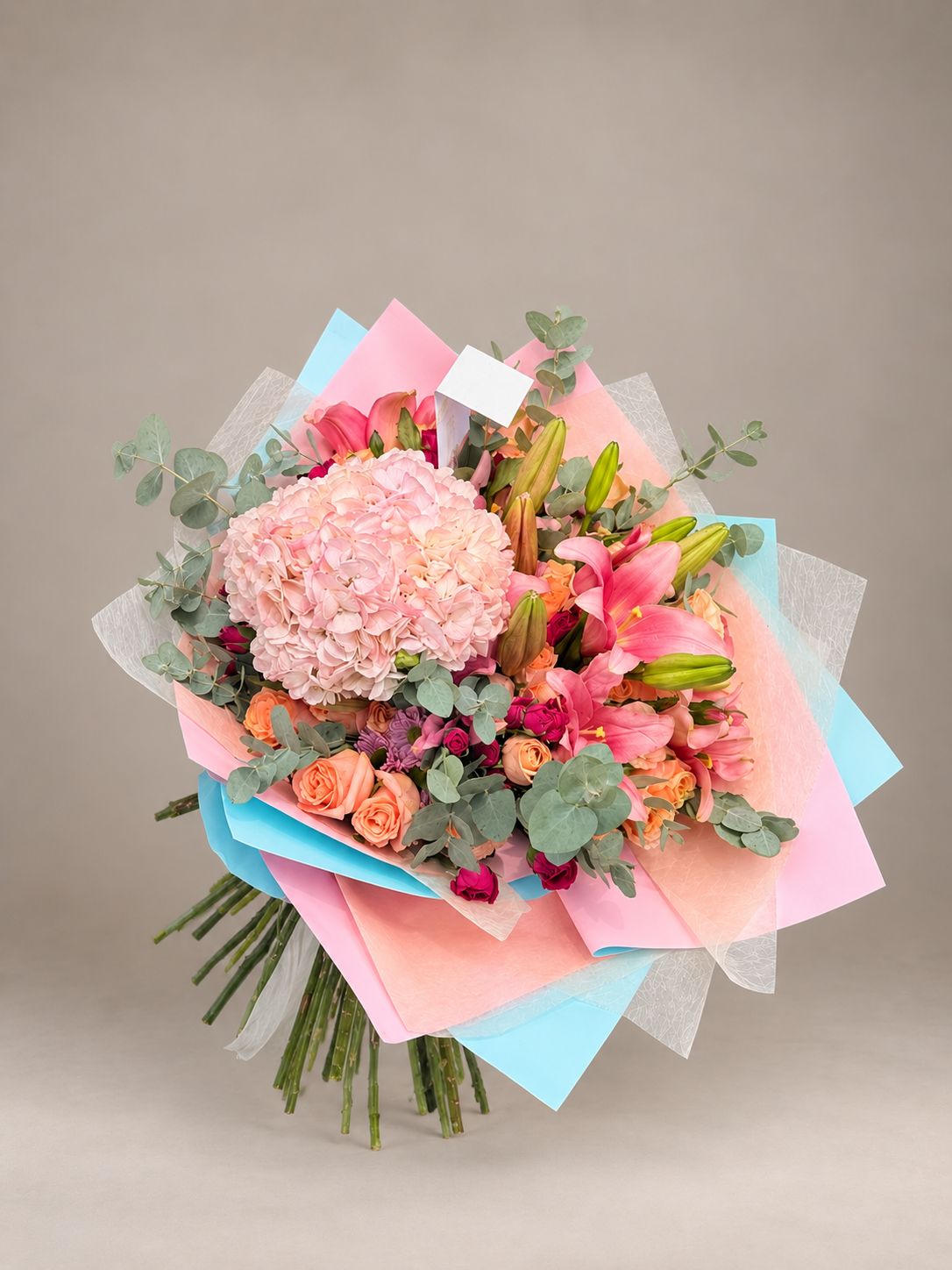 Ramo abundante con hortensia rosada, lirios, rosas naranjas y eucalipto, en papeles rosa y azul.