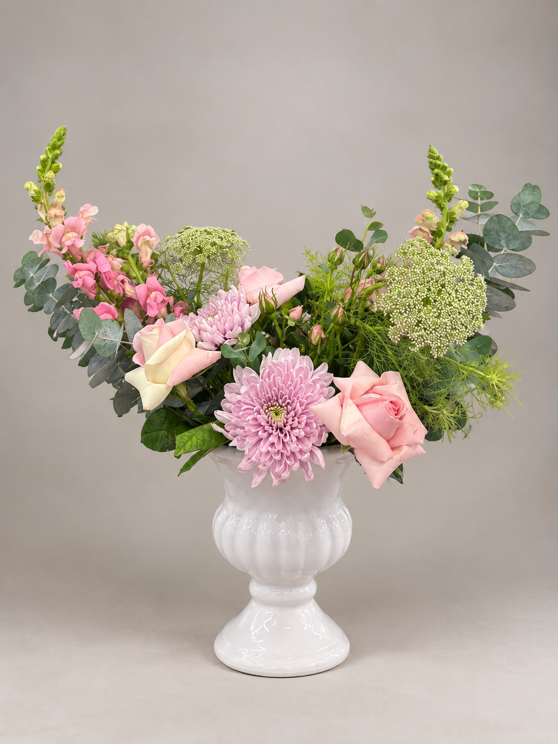Composición suave con rosas, crisantemos, flores rosadas y follaje en una base blanca.